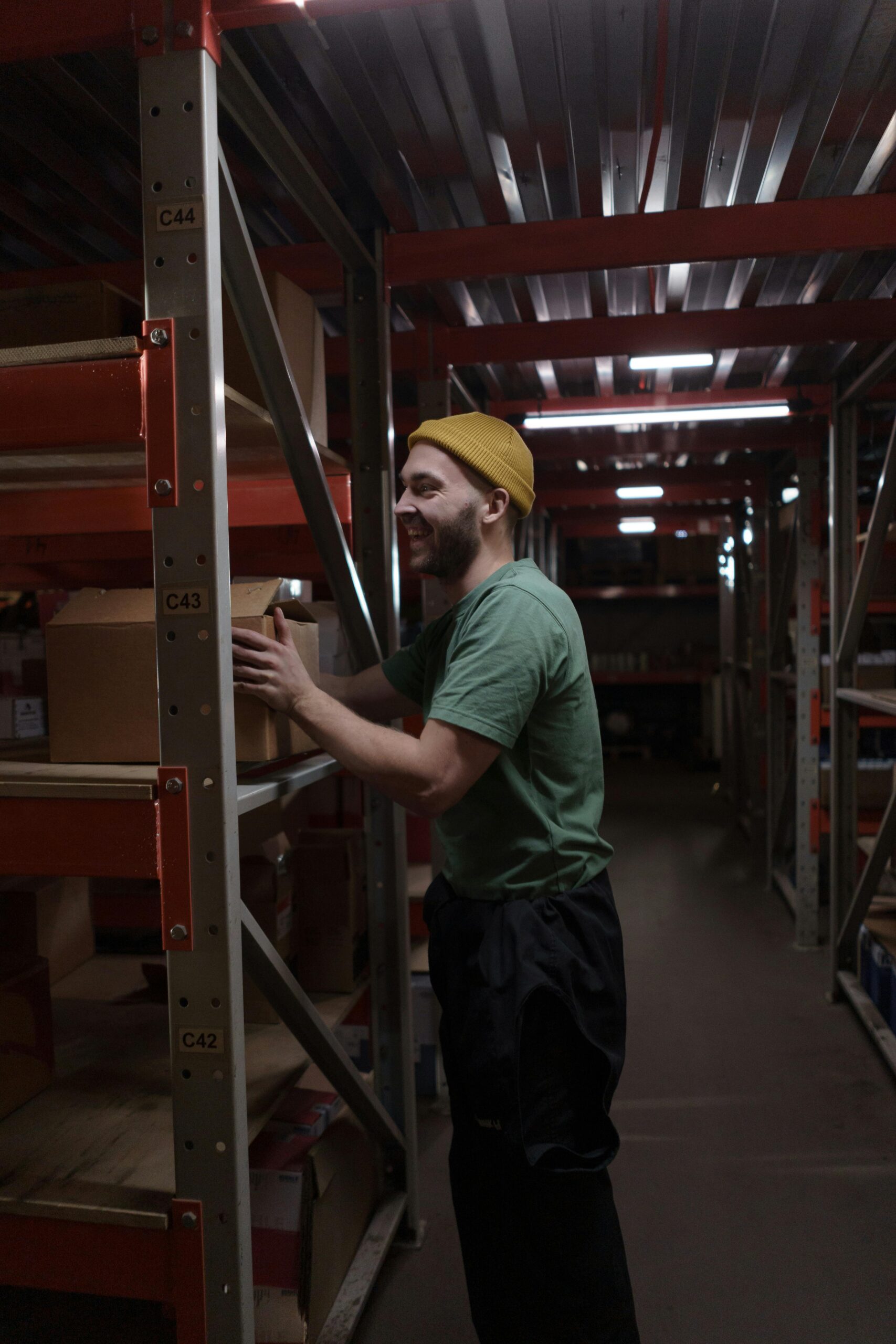 A smiling warehouse employee in a green shirt arranging boxes on shelves in a dimly lit storeroom.